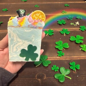 May include: A bar of soap with a St. Patrick's Day theme. The soap is light blue with a green shamrock design. It is topped with colorful swirls, a gold coin, and a small black hat. Green shamrock decorations are scattered around the soap.