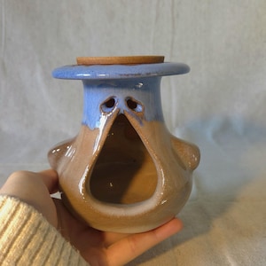 May include: A handmade ceramic bird-shaped incense holder. The holder features a blue top, a brown body, and a wooden lid. The design includes two eye-shaped openings and a large opening in the front. The holder is held in a person's hand.