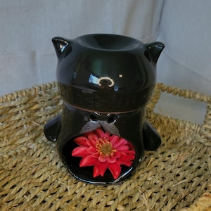 May include: Black ceramic cat-shaped oil burner with a red flower inside. The burner is sitting on a woven basket.