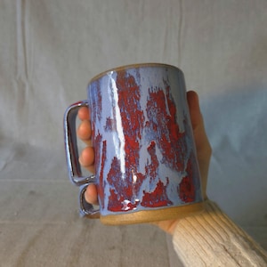 Peut inclure: Une tasse en céramique faite à la main avec un glaçage unique. La tasse présente une base bleu clair avec des stries rouges vives, un bord marron et une anse assortie. La tasse est tenue par une personne.