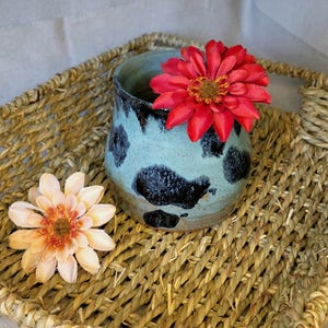 May include: A handmade ceramic vase with a light blue glaze and black spots, holding a red flower. A peach-colored flower rests beside the vase on a woven basket. The vase is a unique decorative piece.