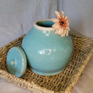 May include: A teal ceramic jar with a lid, sitting on a woven basket. A single peach-colored flower is placed on the top of the jar.