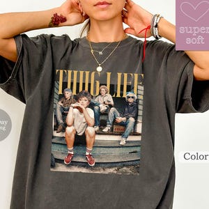 May include: A gray oversized t-shirt with a vintage-style graphic print of a group of people on a porch. The graphic says "THUG LIFE" in bold yellow letters.