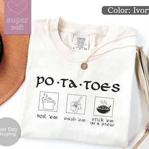 May include: Ivory-colored t-shirt with the word "po-ta-toes" in a whimsical font. Below the text are three illustrated squares with the phrases "boil 'em", "wash 'em", and "stick 'em in a stew". A gold necklace rests on the shirt.