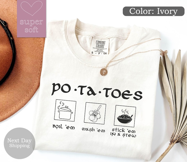 May include: Ivory-colored t-shirt with the word "po-ta-toes" in a whimsical font. Below the text are three illustrated squares with the phrases "boil 'em", "wash 'em", and "stick 'em in a stew". A gold necklace rests on the shirt.