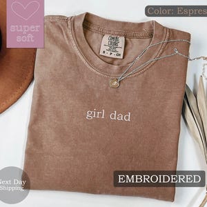 K&ouml;nnte beinhalten: Espresso-braunes T-Shirt mit dem Text "girl dad" in wei&szlig; gestickt. Das Shirt ist aus weichem, bequemem Stoff und hat eine lockere Passform.