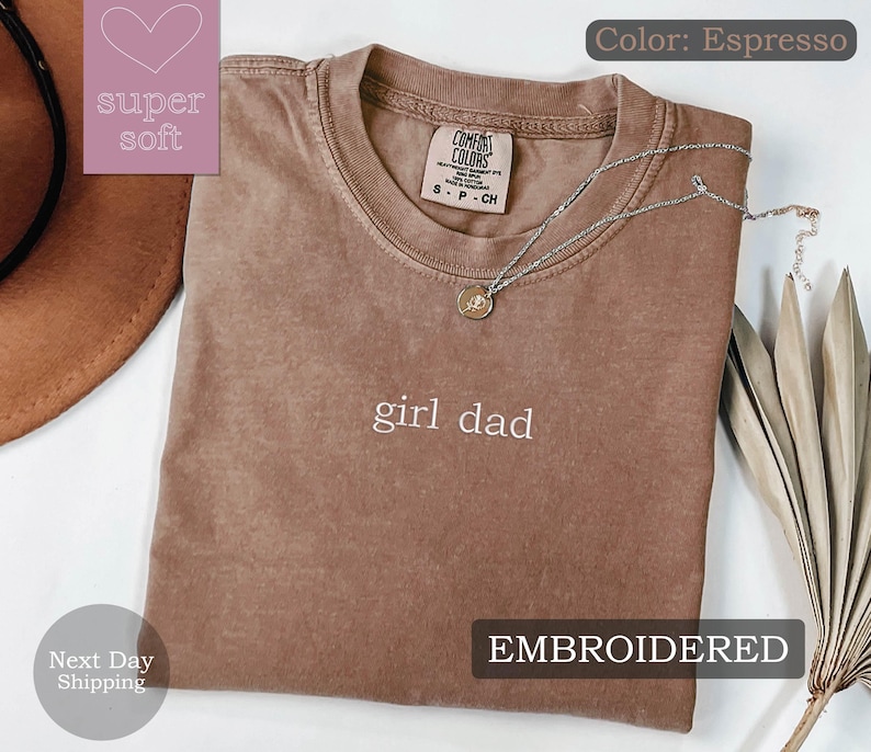 K&ouml;nnte beinhalten: Espresso-braunes T-Shirt mit dem Text "girl dad" in wei&szlig; gestickt. Das Shirt ist aus weichem, bequemem Stoff und hat eine lockere Passform.