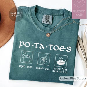 May include: Blue Spruce t-shirt with the word "potatoes" and cooking instructions. The shirt features illustrations of boiling, mashing, and stewing potatoes. A gold necklace with a pendant is on the shirt.