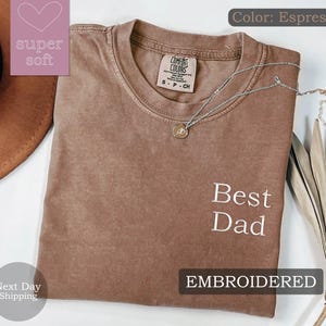 K&ouml;nnte beinhalten: Espresso-braunes T-Shirt mit dem gestickten Text "Best Dad" in Wei&szlig;. Das Shirt ist aus weichem, bequemem Stoff und hat eine lockere Passform.