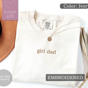K&ouml;nnte beinhalten: Ein T-Shirt in Elfenbein mit dem Text "girl dad", der in braunem Garn gestickt ist. Das Shirt ist gefaltet und liegt flach auf einer wei&szlig;en Oberfl&auml;che. Der Text "EMBROIDERED" ist in Schwarz auf wei&szlig;em Hintergrund gedruckt.