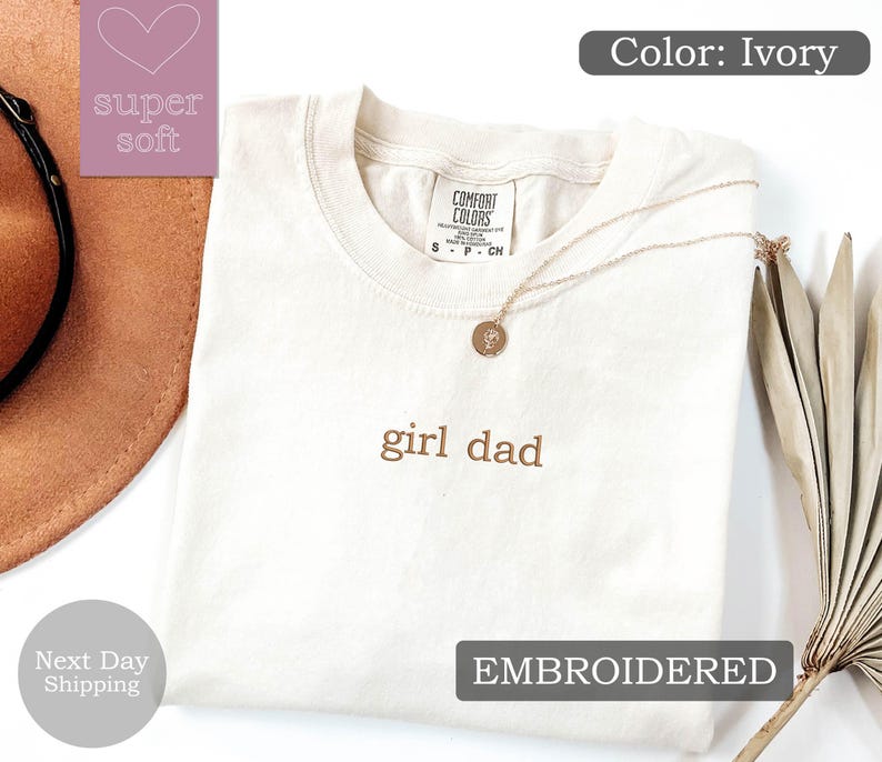 K&ouml;nnte beinhalten: Ein T-Shirt in Elfenbein mit dem Text "girl dad", der in braunem Garn gestickt ist. Das Shirt ist gefaltet und liegt flach auf einer wei&szlig;en Oberfl&auml;che. Der Text "EMBROIDERED" ist in Schwarz auf wei&szlig;em Hintergrund gedruckt.