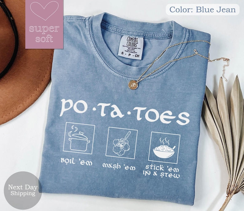 May include: Blue jean colored t-shirt with the word "potatoes" in white, along with illustrated instructions: boil 'em, mash 'em, and stick 'em in a stew. A gold necklace with a pendant is draped on the shirt.