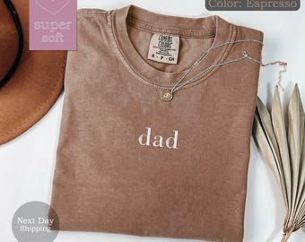 May include: A brown t-shirt with the word "dad" embroidered in white. The shirt is made of a soft, comfortable fabric and has a relaxed fit.