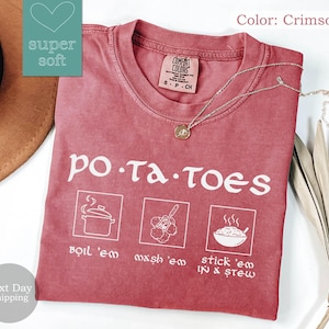 May include: Crimson-colored t-shirt with the word "potatoes" in white, along with three illustrated cooking steps: boil 'em, mash 'em, and stick 'em in stew. The shirt has a crew neck and a small tag on the collar.