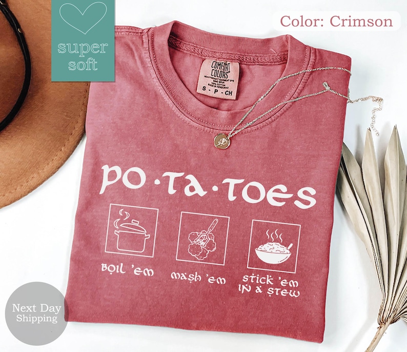 May include: Crimson-colored t-shirt with the word "potatoes" in white, along with three illustrated cooking steps: boil 'em, mash 'em, and stick 'em in stew. The shirt has a crew neck and a small tag on the collar.