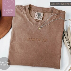 May include: A brown t-shirt with the text "DADDY AF" embroidered on the front. The shirt is made of a soft, comfortable fabric and has a relaxed fit.