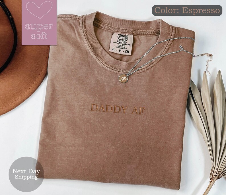May include: A brown t-shirt with the text "DADDY AF" embroidered on the front. The shirt is made of a soft, comfortable fabric and has a relaxed fit.