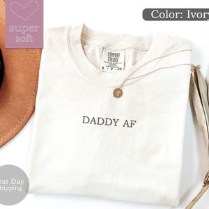 May include: Ivory t-shirt with the text "DADDY AF" printed on the front. The shirt is folded and laid flat on a white surface.