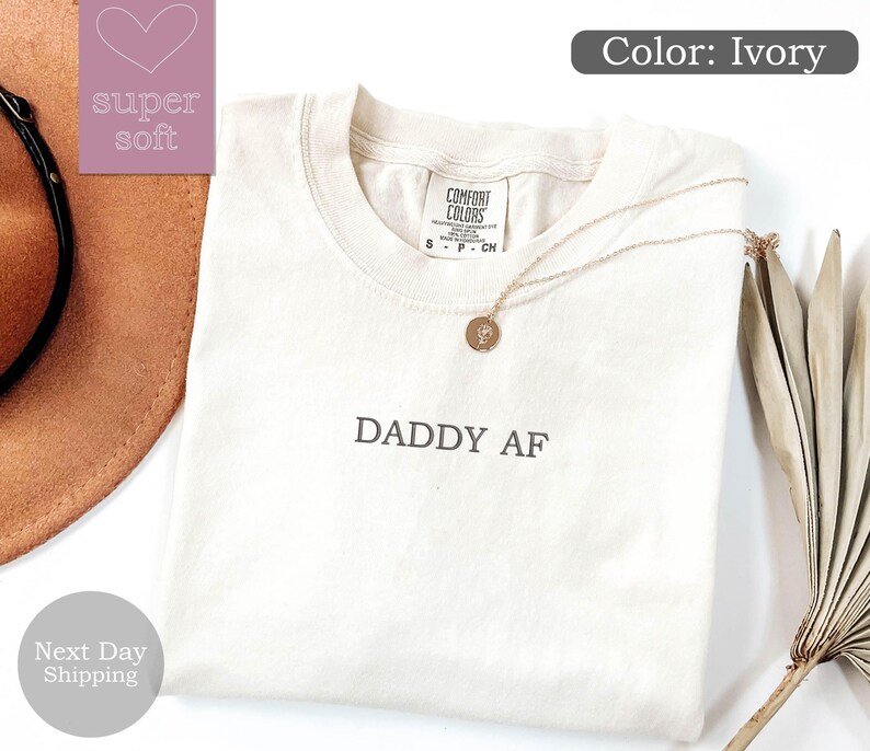 May include: Ivory t-shirt with the text "DADDY AF" printed on the front. The shirt is folded and laid flat on a white surface.