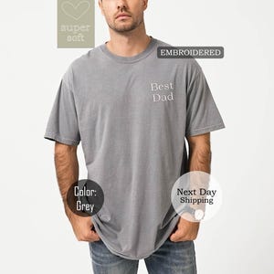 K&ouml;nnte beinhalten: Graues T-Shirt mit dem gestickten Text "Best Dad" auf der Vorderseite. Das Shirt ist aus weichem, bequemem Stoff.