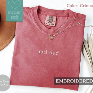K&ouml;nnte beinhalten: Ein T-Shirt in Rot mit der Aufschrift "girl dad", die in Wei&szlig; gestickt ist. Das Shirt ist aus einem weichen, bequemen Stoff.