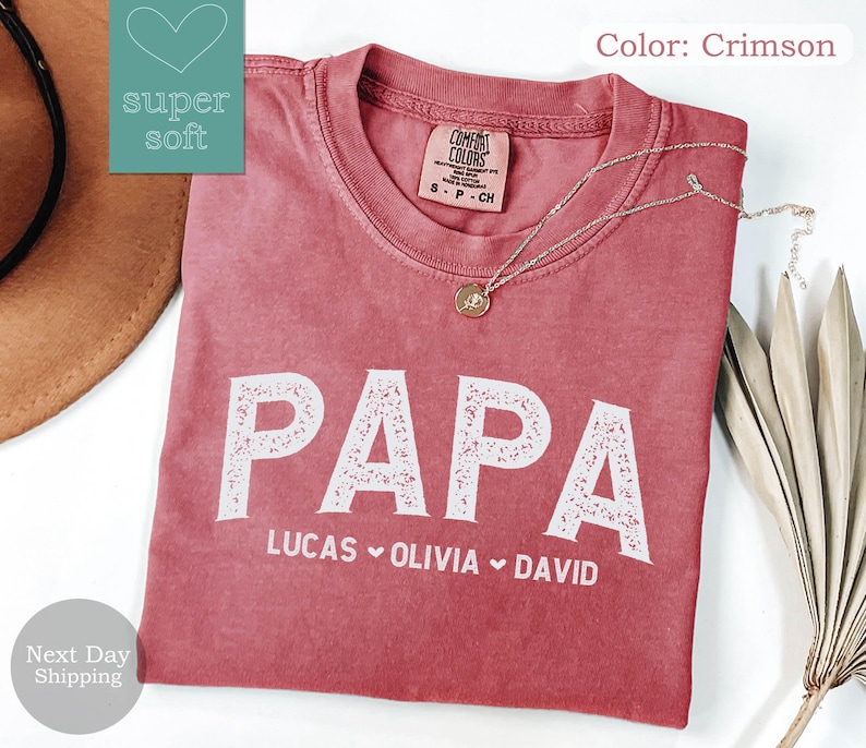 May include: Crimson red t-shirt with the word "PAPA" in white with a decorative floral design. The names "LUCAS", "OLIVIA", and "DAVID" are written below in white.