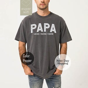 May include: A dark gray t-shirt with the word "PAPA" in white lettering. The names "Lucas", "Olivia", and "David" are printed below the word "PAPA".