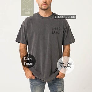 K&ouml;nnte beinhalten: Ein graues T-Shirt mit kurzen &Auml;rmeln und dem gestickten Text "Best Dad" auf der Vorderseite. Das Shirt ist aus weichem, bequemem Stoff.