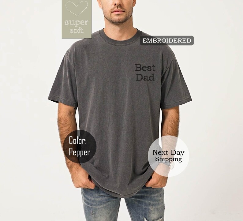 K&ouml;nnte beinhalten: Ein graues T-Shirt mit kurzen &Auml;rmeln und dem gestickten Text "Best Dad" auf der Vorderseite. Das Shirt ist aus weichem, bequemem Stoff.