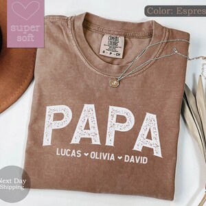 May include: Espresso brown t-shirt with the word "PAPA" in white with a decorative floral design. Below the word "PAPA" are the names "LUCAS", "OLIVIA", and "DAVID" separated by hearts.