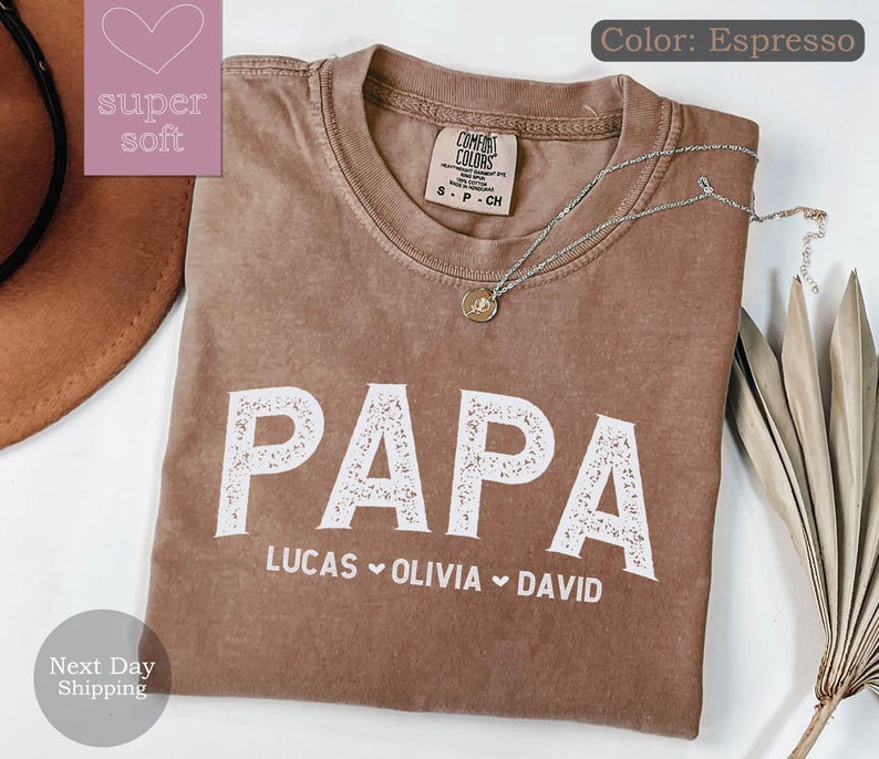 May include: Espresso brown t-shirt with the word "PAPA" in white with a decorative floral design. Below the word "PAPA" are the names "LUCAS", "OLIVIA", and "DAVID" separated by hearts.