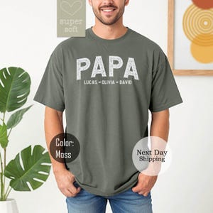 May include: A green t-shirt with the word "PAPA" in white text. Below the word "PAPA" is the text "LUCAS OLIVIA DAVID" in smaller white text.