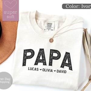 May include: Ivory t-shirt with the word "PAPA" in black with a distressed texture. Below the word "PAPA" are the names "LUCAS", "OLIVIA", and "DAVID" separated by hearts.