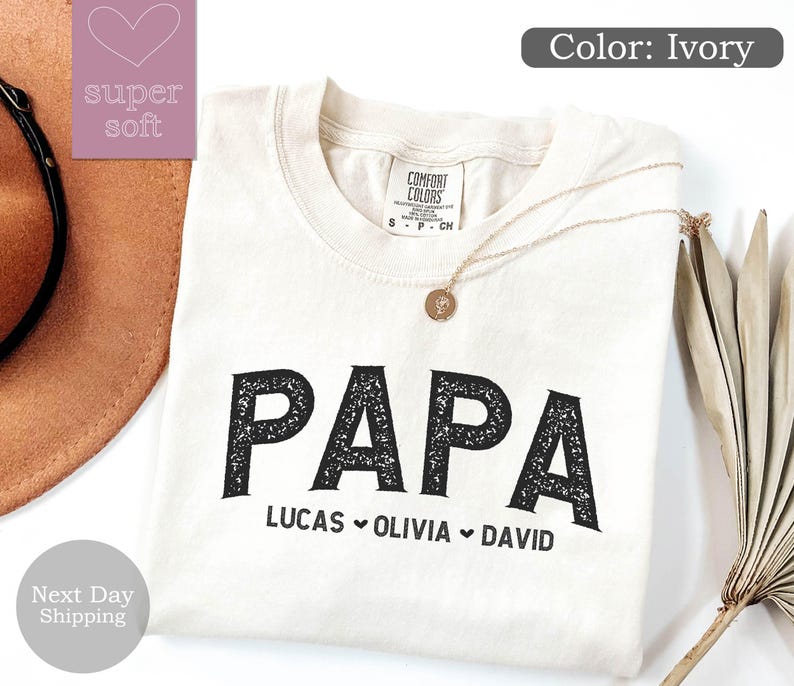 May include: Ivory t-shirt with the word "PAPA" in black with a distressed texture. Below the word "PAPA" are the names "LUCAS", "OLIVIA", and "DAVID" separated by hearts.