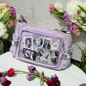 May include: A lavender-colored shoulder bag with clear plastic panels. The panels feature various anime character stickers and photos. The bag has a zipper closure and adjustable straps.