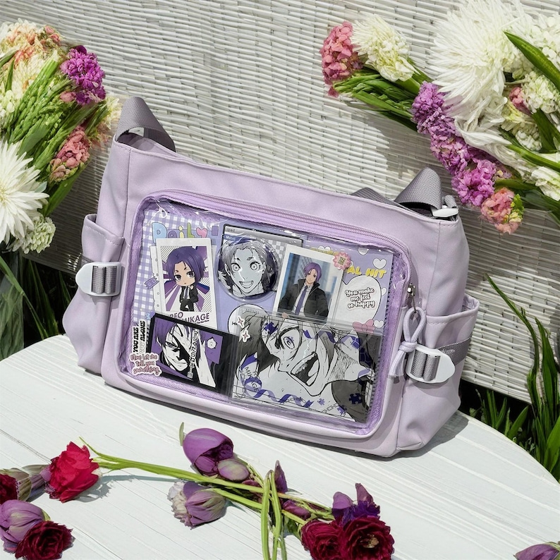 May include: A lavender-colored shoulder bag with clear plastic panels. The panels feature various anime character stickers and photos. The bag has a zipper closure and adjustable straps.
