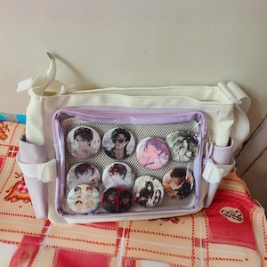 May include: A white and purple messenger bag with a clear plastic pocket on the front. The pocket contains a variety of round pins with images of people on them.