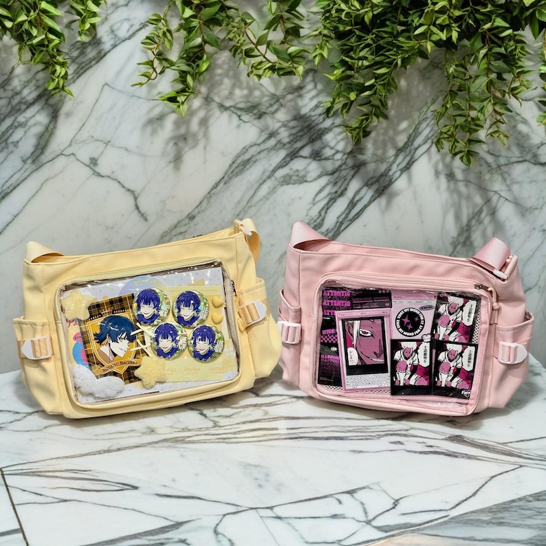 May include: Two canvas shoulder bags with clear plastic front pockets. The yellow bag has anime character pins inside. The pink bag has black and white graphic stickers inside. Both bags have adjustable straps.