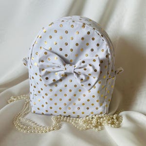 May include: White backpack with gold polka dots and a large bow. The backpack has a rounded shape and small handles. A strand of pearls is draped in front of the backpack.