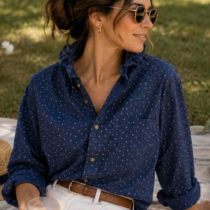 May include: A navy blue button-down shirt with white polka dots, paired with white pants and a brown belt. A straw hat, picnic basket, and food items are arranged on a blanket. The scene is set outdoors on a sunny day.