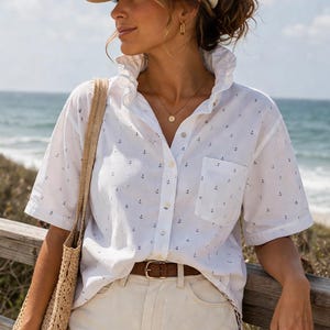 May include: A white button-down shirt with small blue anchor patterns, paired with light-colored denim shorts and a beige baseball cap. A brown belt and a woven tote bag complete the casual, summery outfit. The background shows the ocean and a wooden fence.