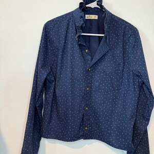 May include: A navy blue button-down shirt with a ruffled collar and long sleeves. The shirt is adorned with a pattern of small white and red shapes. The shirt is hanging on a white hanger.