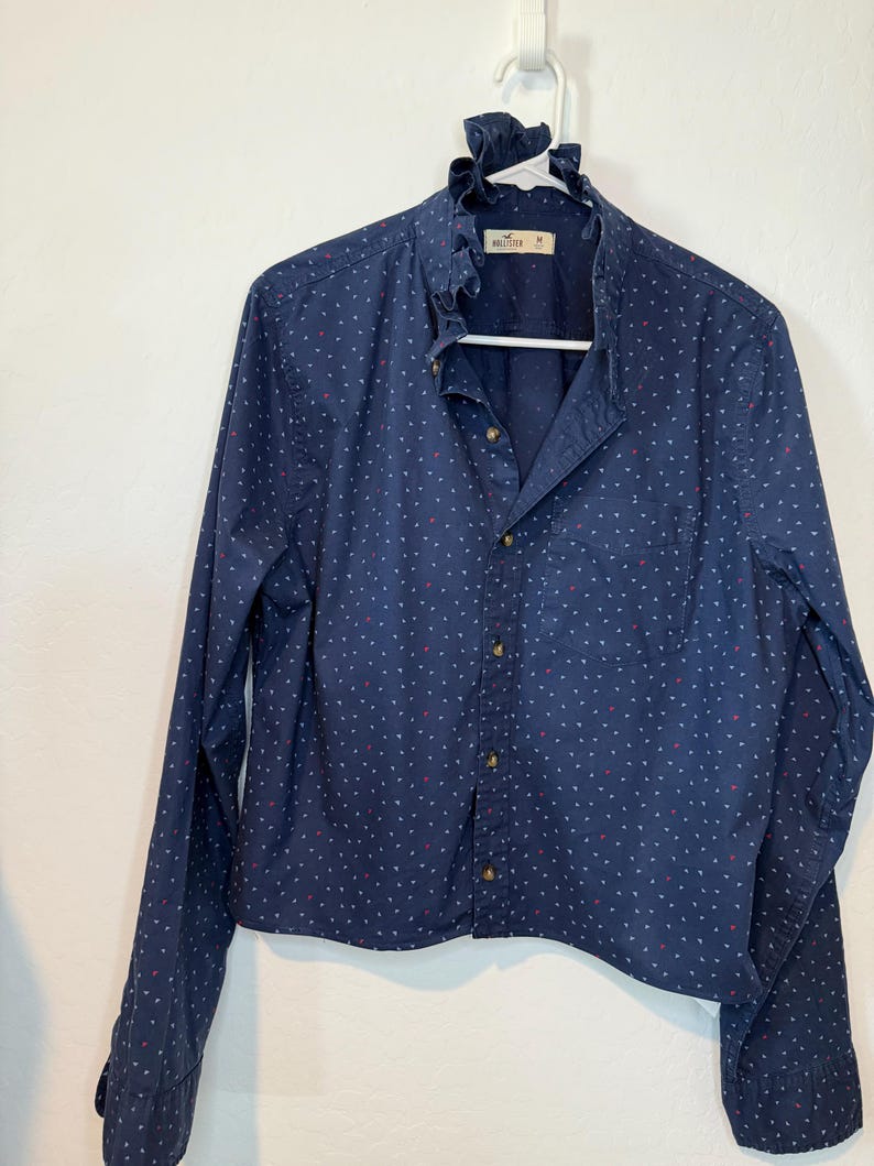May include: A navy blue button-down shirt with a ruffled collar and long sleeves. The shirt is adorned with a pattern of small white and red shapes. The shirt is hanging on a white hanger.