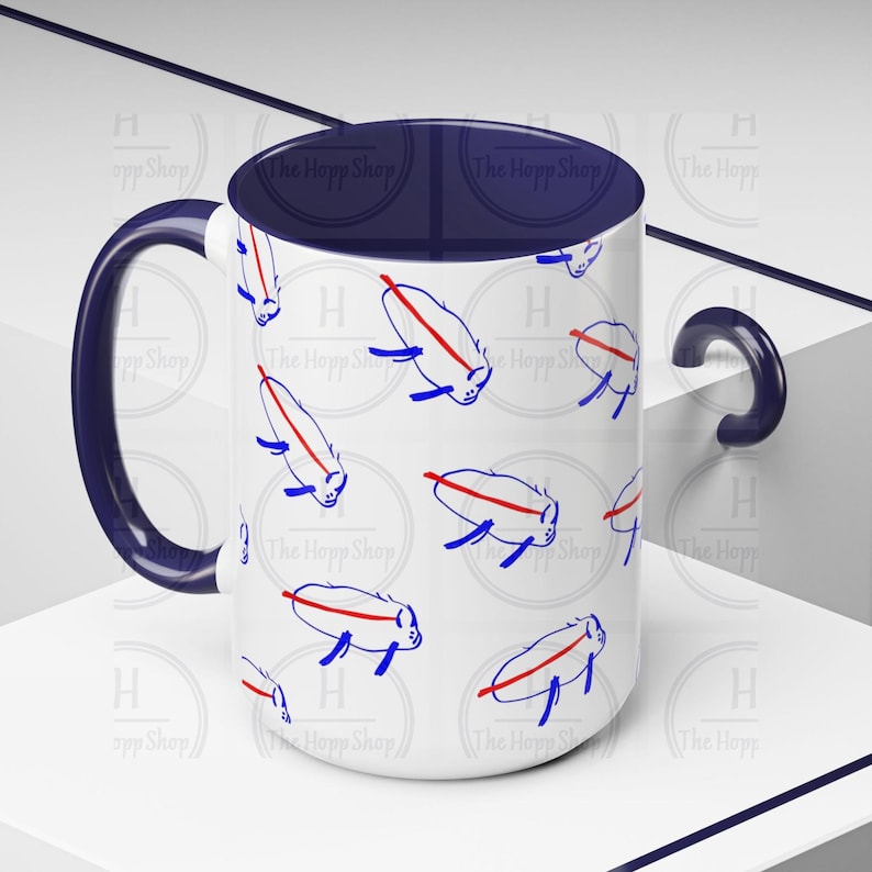 May include: White ceramic mug with a navy blue handle and interior. The mug features a repeating pattern of stylized blue and red creatures. The text "The Hopp Shop" is printed around the base of the mug.