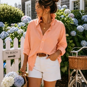 May include: A woman wearing a peach-colored button-down shirt with a ruffled collar and white shorts. She is holding a woven basket filled with blue and white hydrangeas. A brown leather belt and a sign that says "TO BEACH" are also visible.