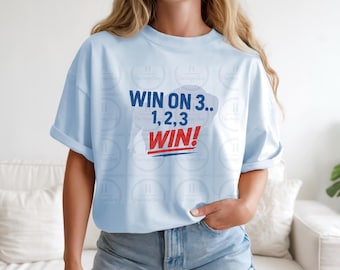 May include: A light blue short-sleeved t-shirt featuring the dark blue and red text "WIN ON 3.. 1, 2, 3 WIN!" printed on the front. The shirt has a crew neckline and a relaxed fit, paired with light blue denim shorts with a cuffed hem.