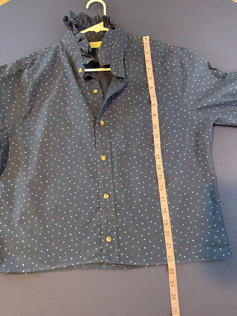 May include: Navy blue button-down shirt with a ruffle collar and small white and red triangle pattern. The shirt has gold-colored buttons and a single chest pocket. A measuring tape shows the shirt is approximately 26 inches long.
