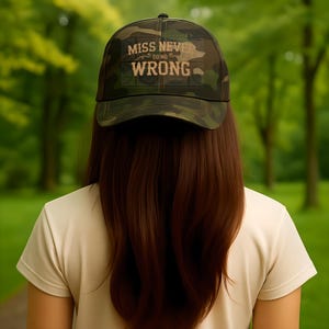 May include: A camouflage baseball cap with the text "MISS NEVER DO NO WRONG" in a gold color. The cap is worn by a person with long, dark brown hair, standing in a park with green trees in the background.
