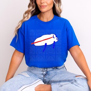 Pu&ograve; includere: Una t-shirt a maniche corte di colore blu reale brillante con una grafica centrale raffigurante una creatura bianca di forma ovale con una linea orizzontale rossa sul corpo e appendici blu. La grafica &egrave; su uno sfondo di quadrati blu.