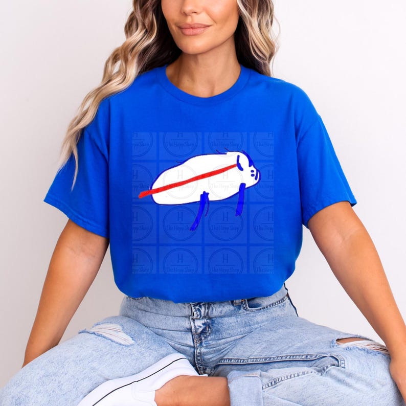 Pu&ograve; includere: Una t-shirt a maniche corte di colore blu reale brillante con una grafica centrale raffigurante una creatura bianca di forma ovale con una linea orizzontale rossa sul corpo e appendici blu. La grafica &egrave; su uno sfondo di quadrati blu.